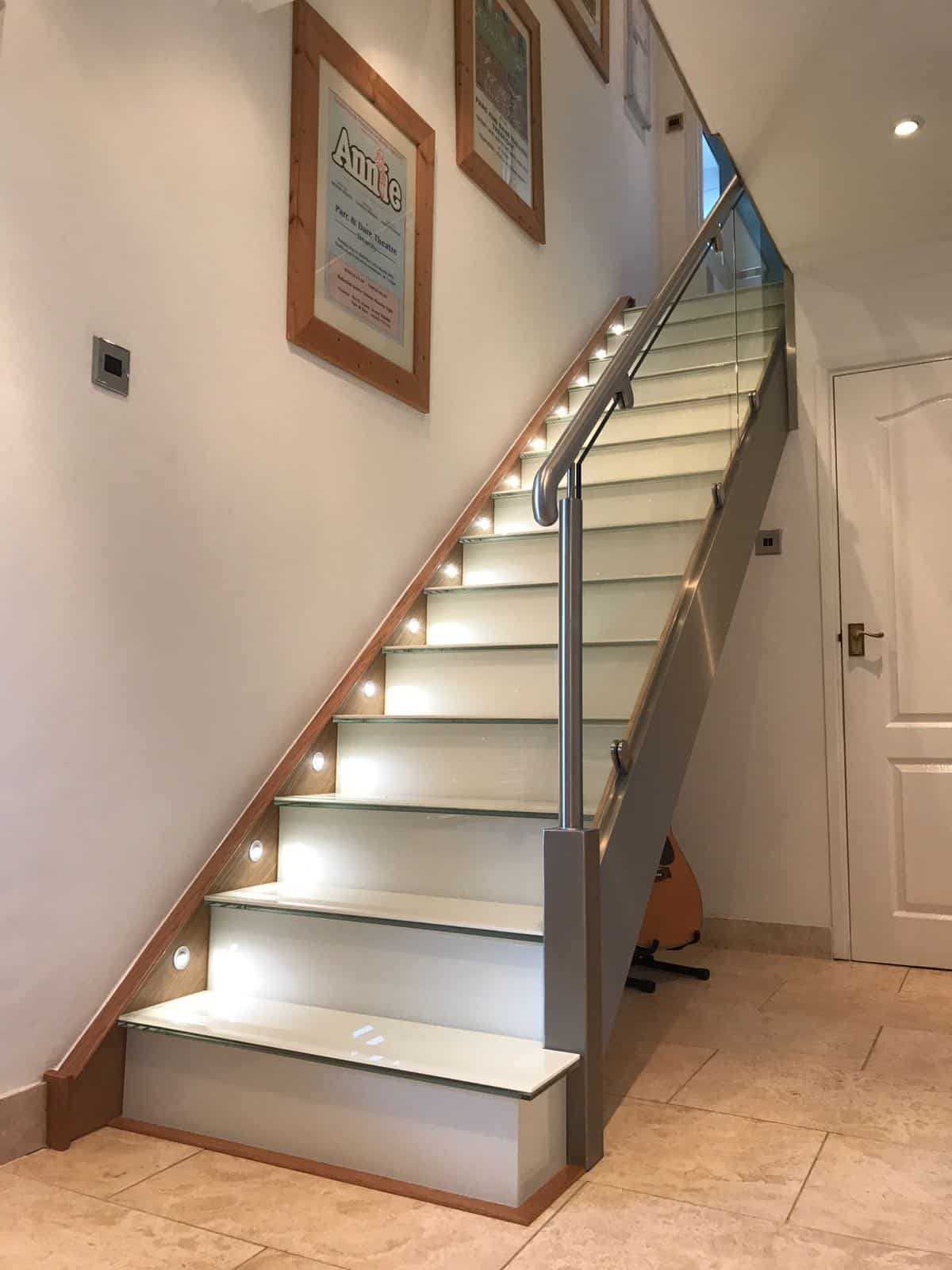 Straight white staircase with grey stringers, recessed step lighting, a glass balustrade and a stainless steel handrail.