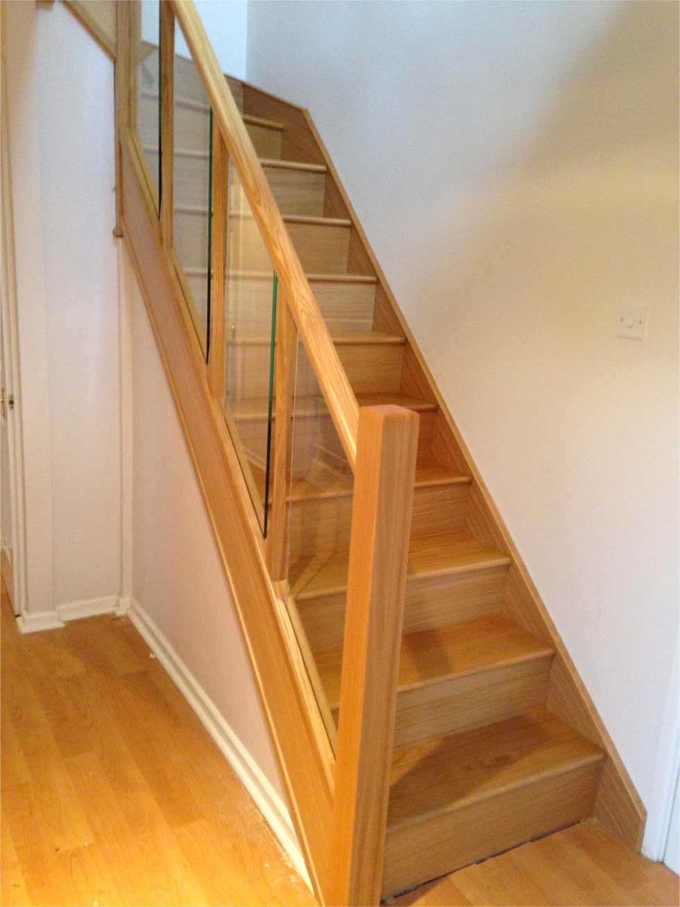Straight oak staircase with closed wooden treads, a square newel post and clear glass panels fitted within the wooden handrail and base rail.