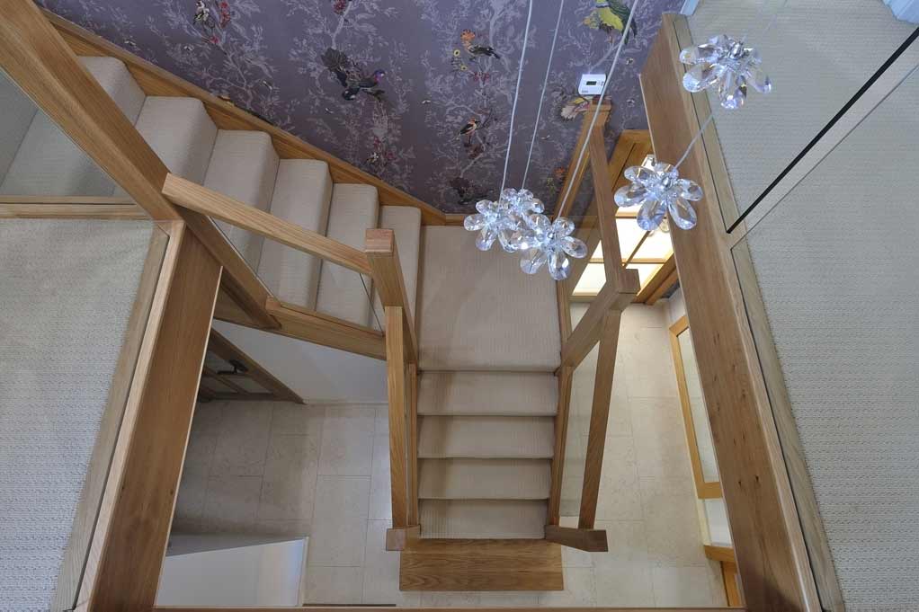 Overhead view of a turning carpeted staircase with oak-framed glass balustrades beneath flower-shaped pendant lights.