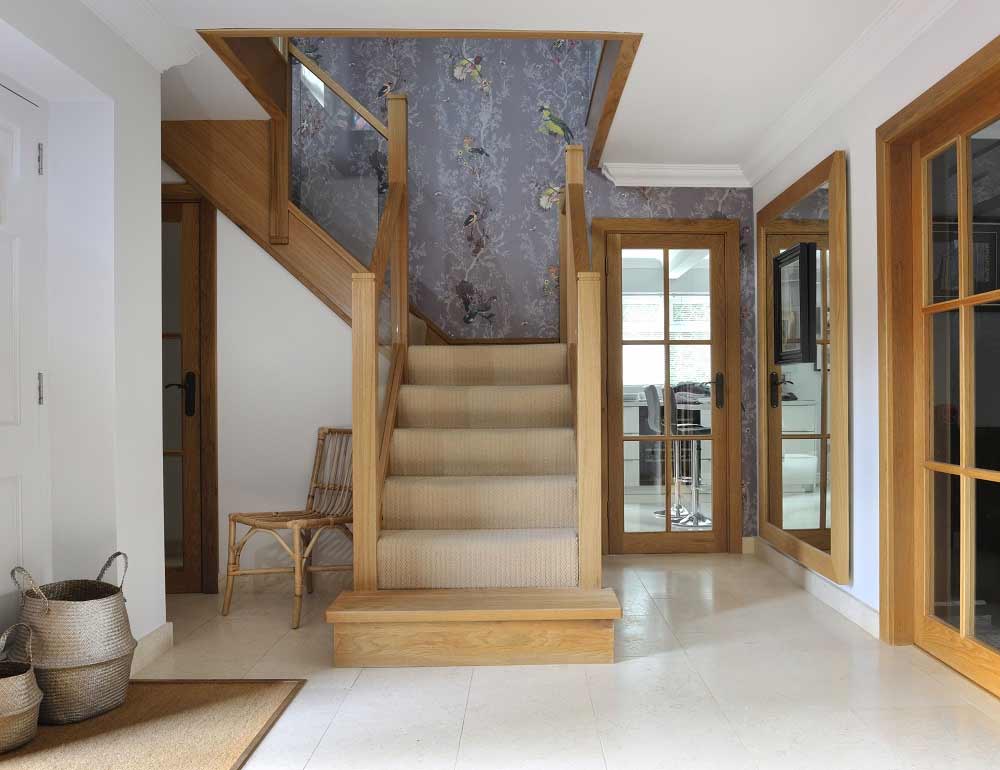 Cream carpeted oak staircase with integrated glass panels and a rectangular wooden feature step in a tiled hallway with floral wallpaper.