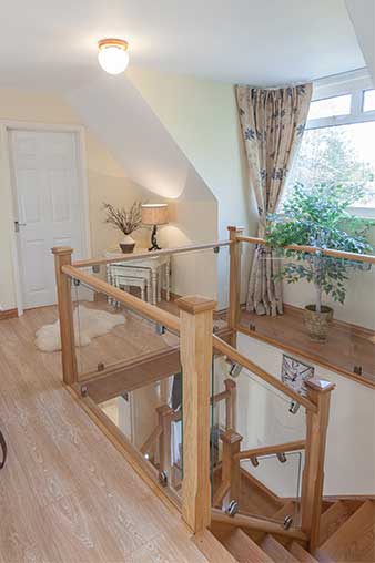 Oak staircase and upper landing with square wooden posts, matching handrails and clear glass panels held by metal brackets beside a large window.