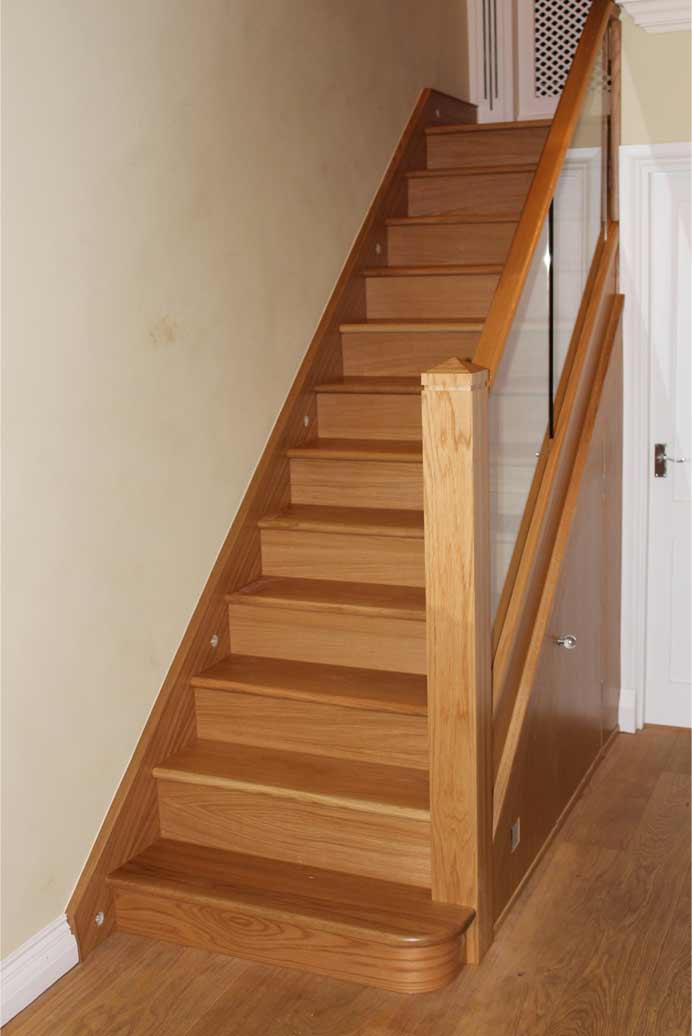 Straight oak staircase with integrated glass balustrades, a square newel post, a rounded bottom step and small recessed lights along the wall-side stringer.