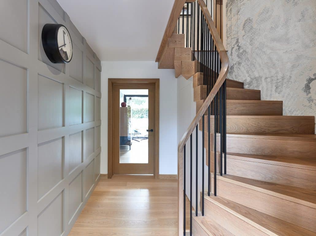 Oak staircase with slender black metal spindles and a curved wooden handrail beside a grey panelled hallway wall.
