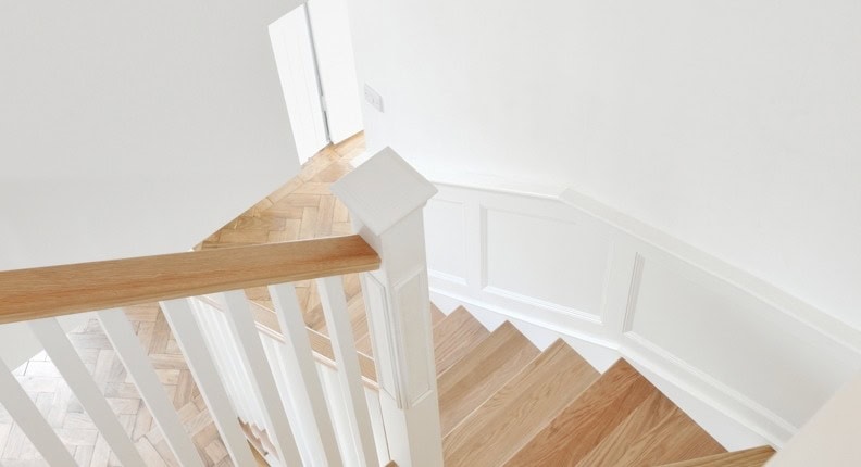 View down natural oak staircase treads beside white square spindles, a white box newel post and an oak handrail in a white panelled stairwell.