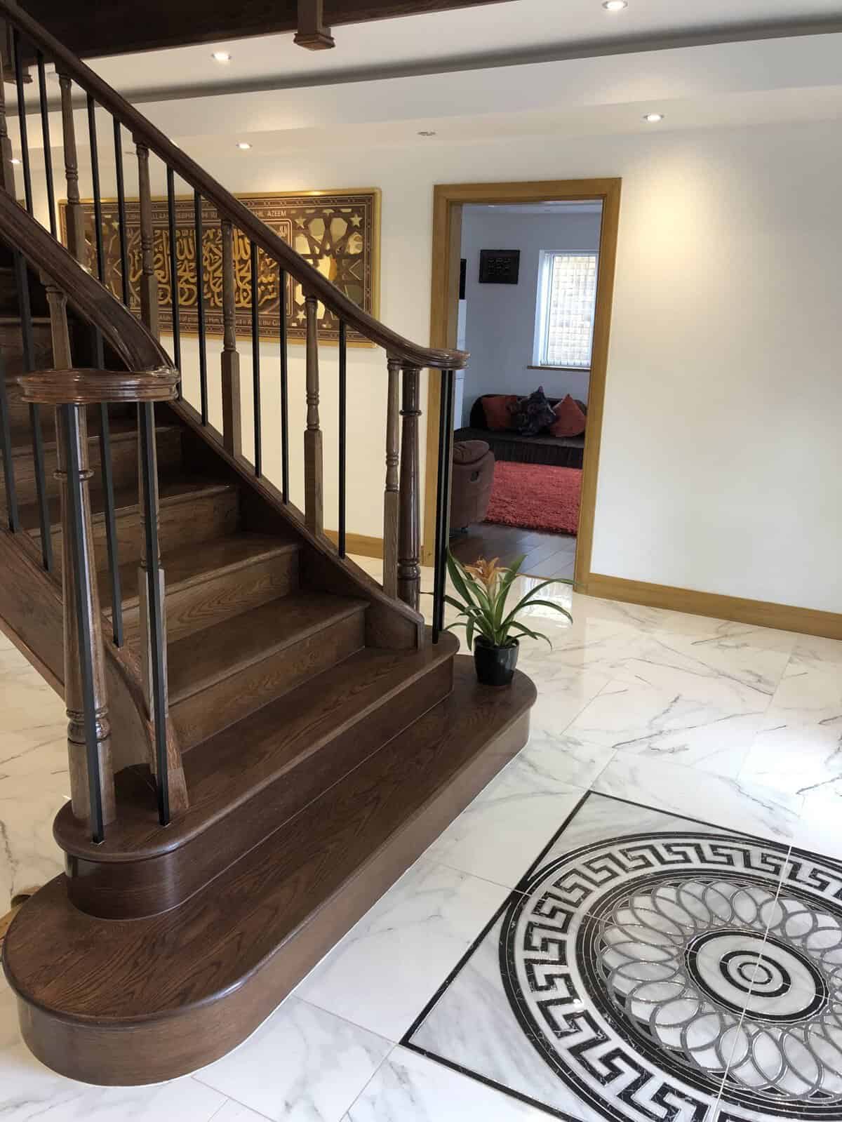 Dark wooden staircase with decorative metal spindles, curved wooden handrails and broad rounded bottom steps in a marble-tiled hallway.