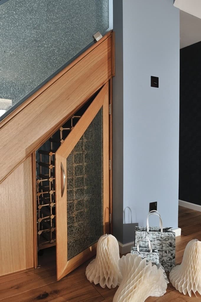 Partly open oak understair cupboard with a patterned glass door revealing fitted wine bottle racks beneath a glass-balustrade staircase.