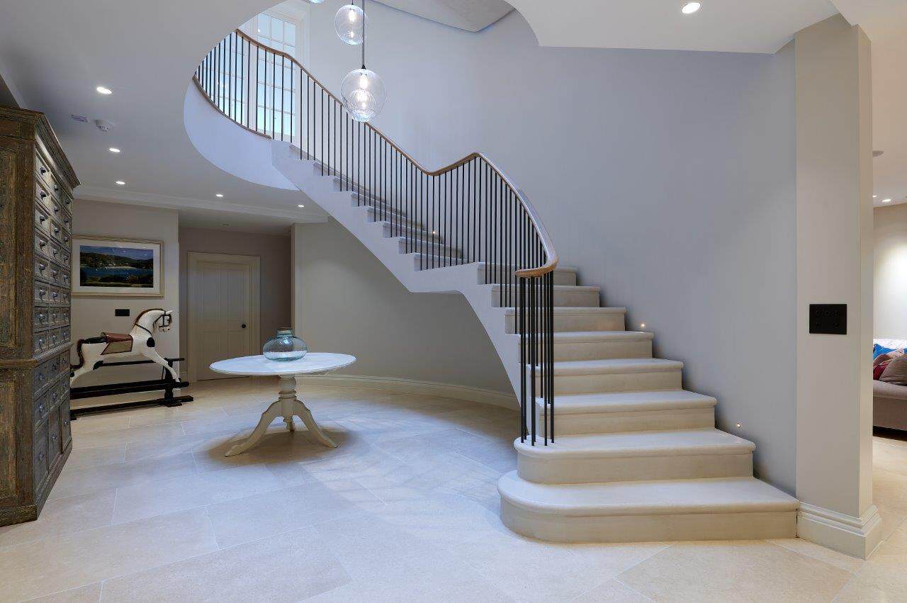 Sweeping pale stone staircase with slender black iron spindles, a continuous curved oak handrail and broad rounded lower steps in a spacious hallway.