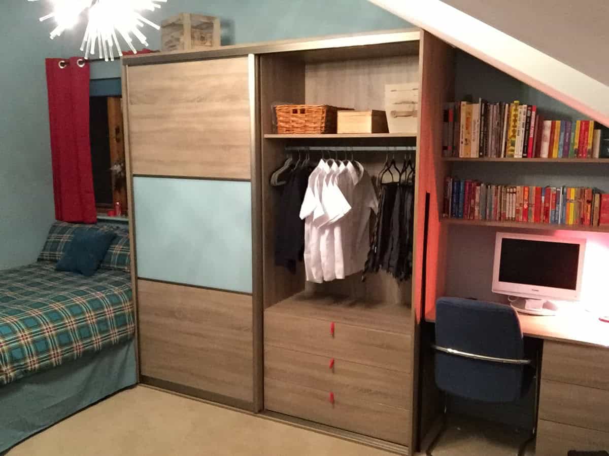 Fitted bedroom wardrobe with a sliding wood-and-blue-panel door, hanging space and drawers beside a built-in desk and bookshelves under a sloping ceiling.