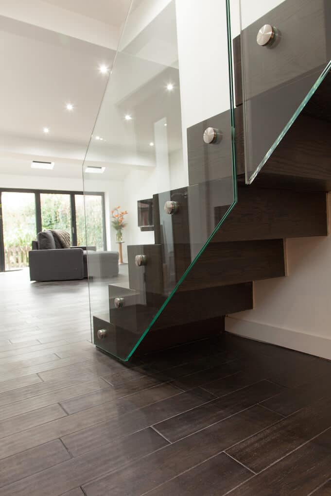 Side view of a dark wooden zigzag staircase with a frameless glass balustrade secured by round metal fixings in an open-plan interior.