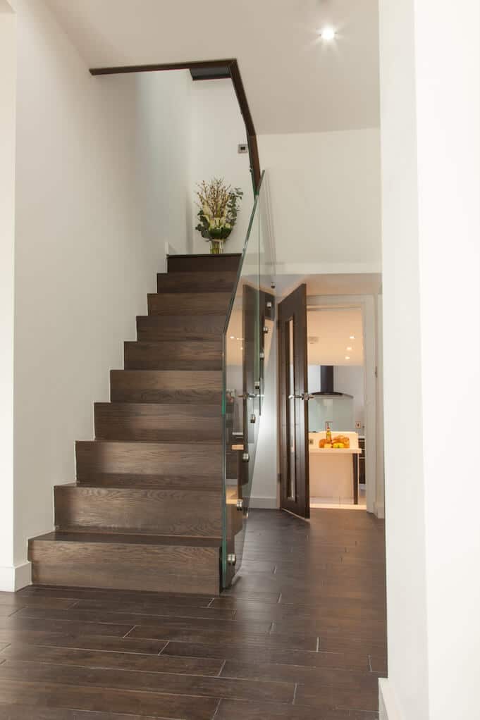 Straight dark wooden staircase with closed risers and a frameless glass balustrade leading to a matching glass-fronted landing.
