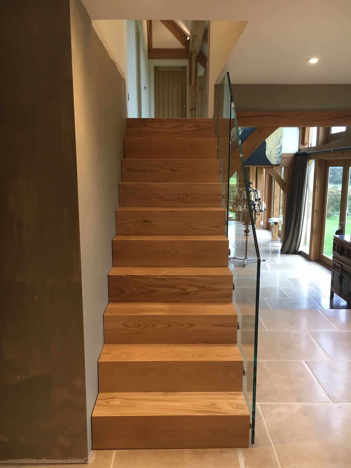 Straight oak staircase with closed risers and a frameless glass balustrade in a timber-framed interior beside full-height windows.