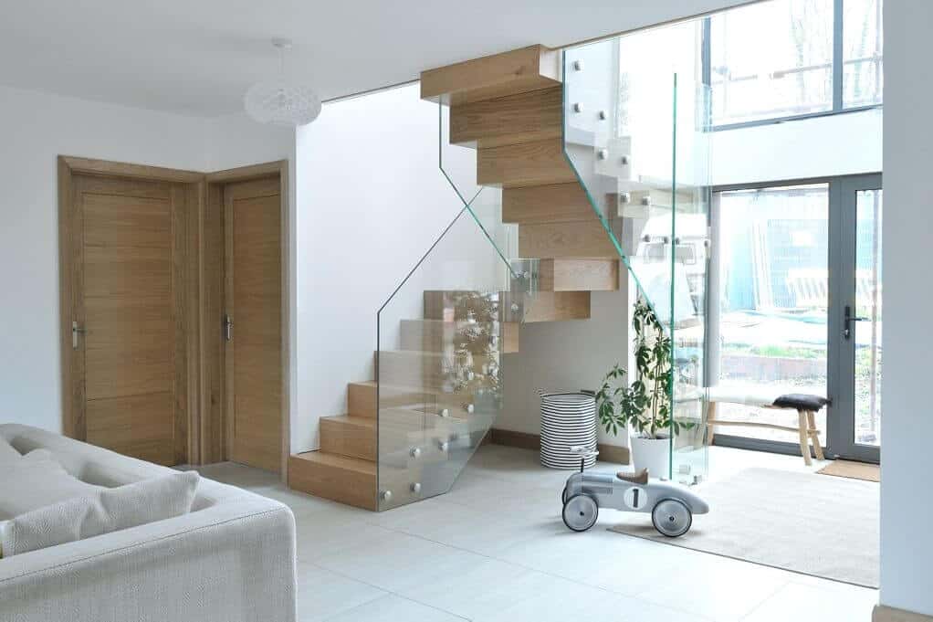 Wooden zigzag staircase with frameless glass balustrades in a bright open-plan room beside full-height windows.