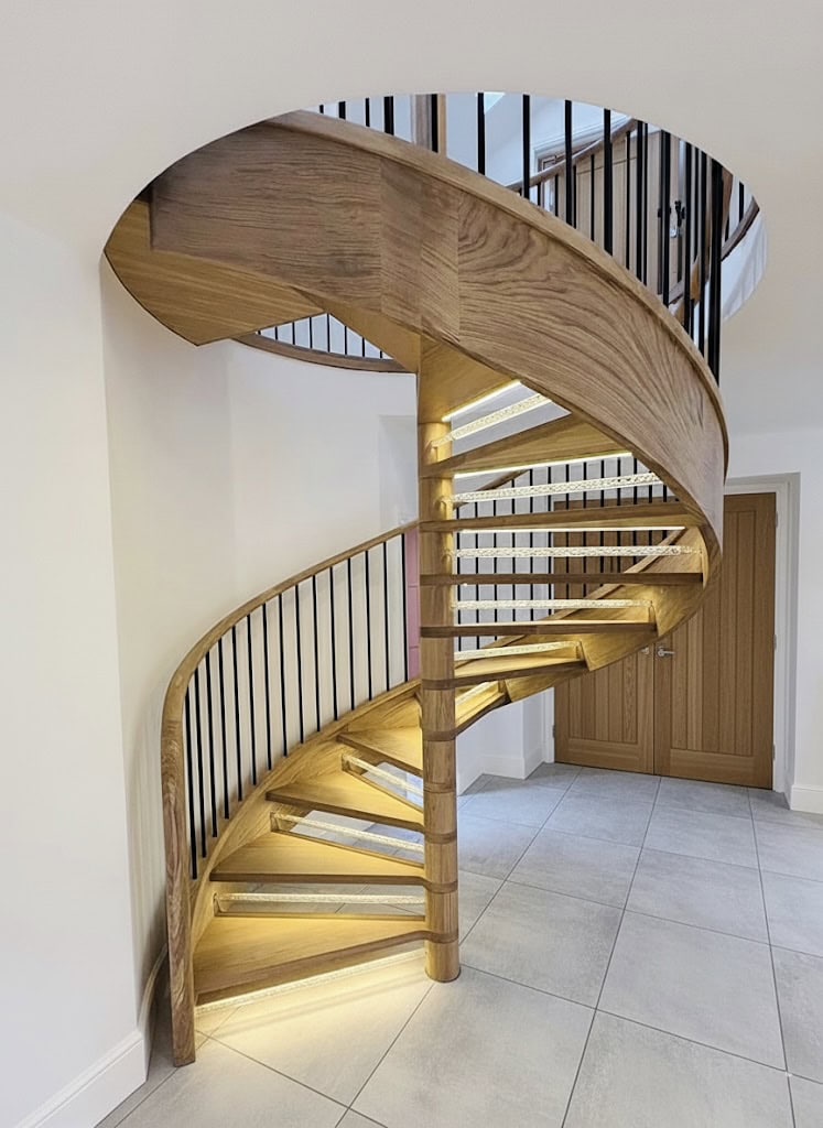 Illuminated oak spiral staircase with open wooden treads, a central timber column, curved oak stringer and slender black metal spindles.