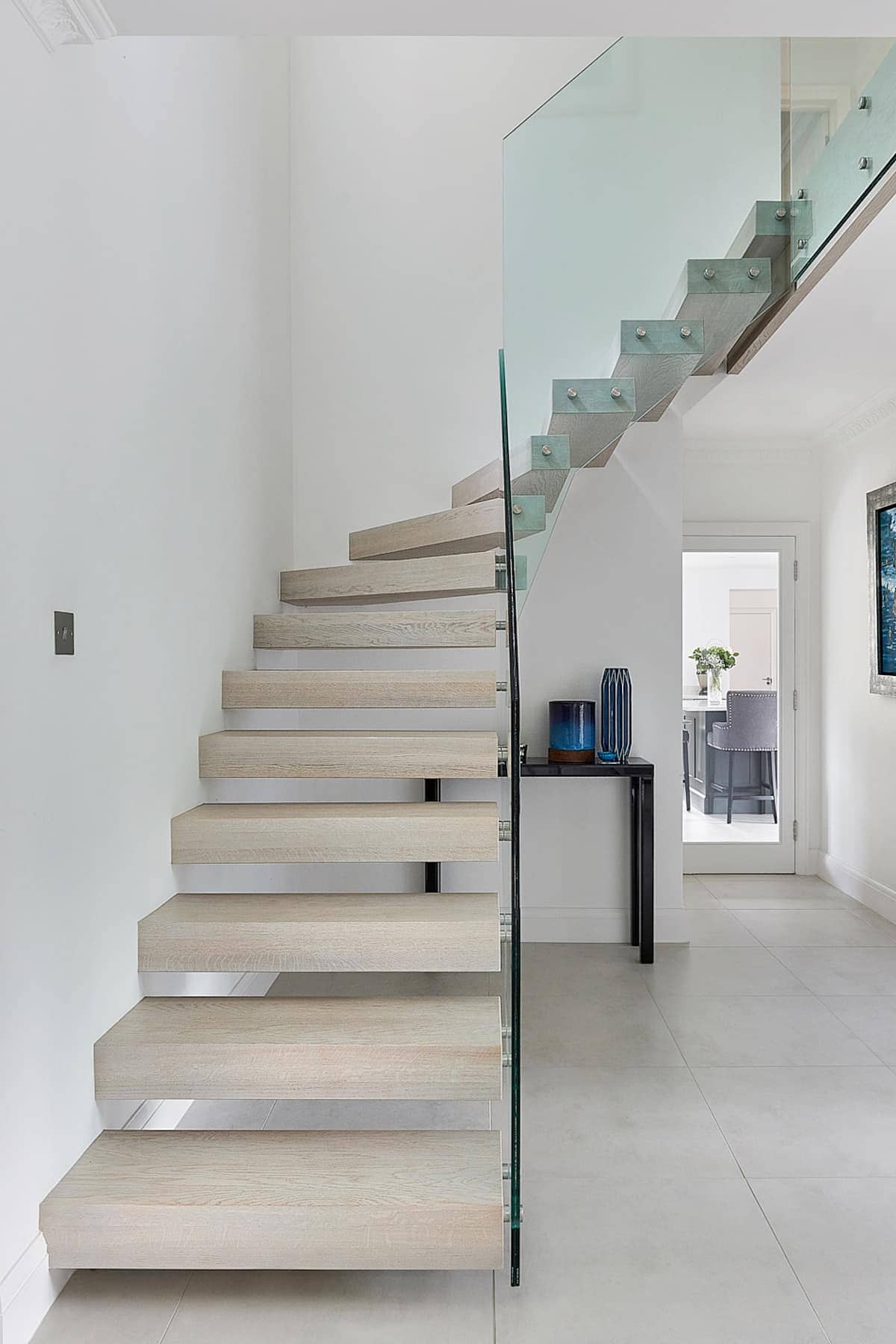 Pale wooden floating staircase with open treads and a frameless glass balustrade in a bright white hallway.