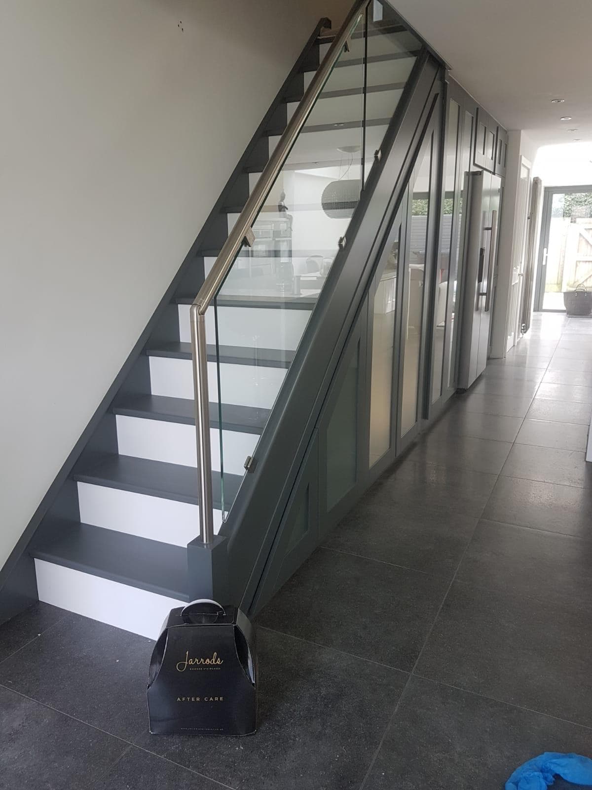 Dark grey staircase with white risers, glass balustrades, a stainless steel handrail and matching fitted cupboards along a dark tiled hallway.