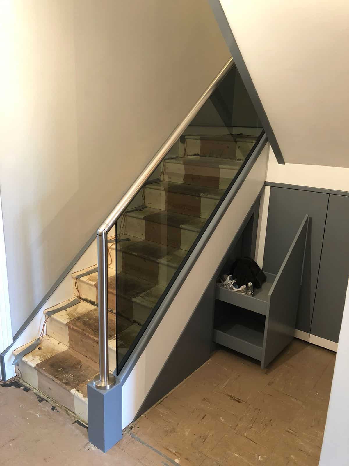 Staircase during renovation with exposed wooden treads, a grey frame, dark glass balustrade and stainless steel handrail beside an open pull-out understair drawer.