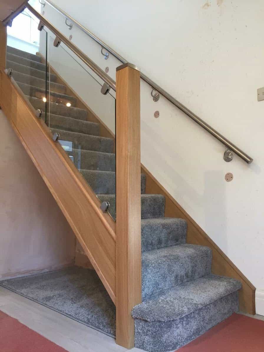 Grey carpeted oak staircase with square wooden newel posts, bracketed glass panels and stainless steel handrails along the glass and opposite wall.