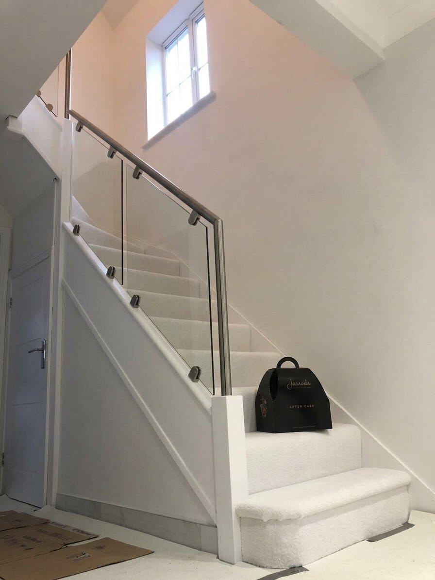 White carpeted staircase with white timber framing, clear clamped glass panels and a stainless steel handrail beneath a high hallway window.