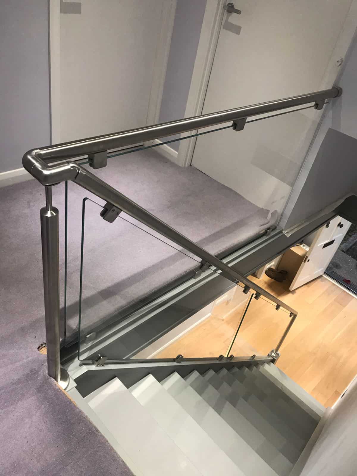 View down a grey staircase beside a carpeted landing with clear glass panels, stainless steel posts and a continuous curved metal handrail.
