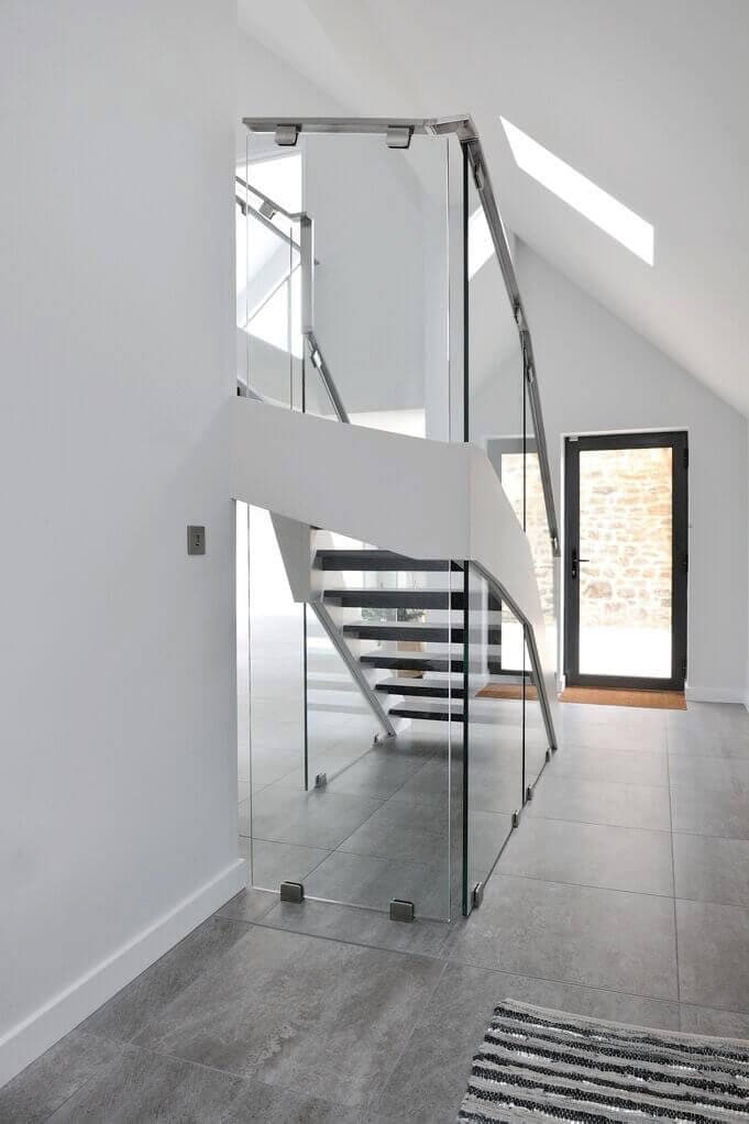 View along a glass balustrade towards an open-riser staircase with dark treads, white stringers and polished metal handrails in a white hallway.