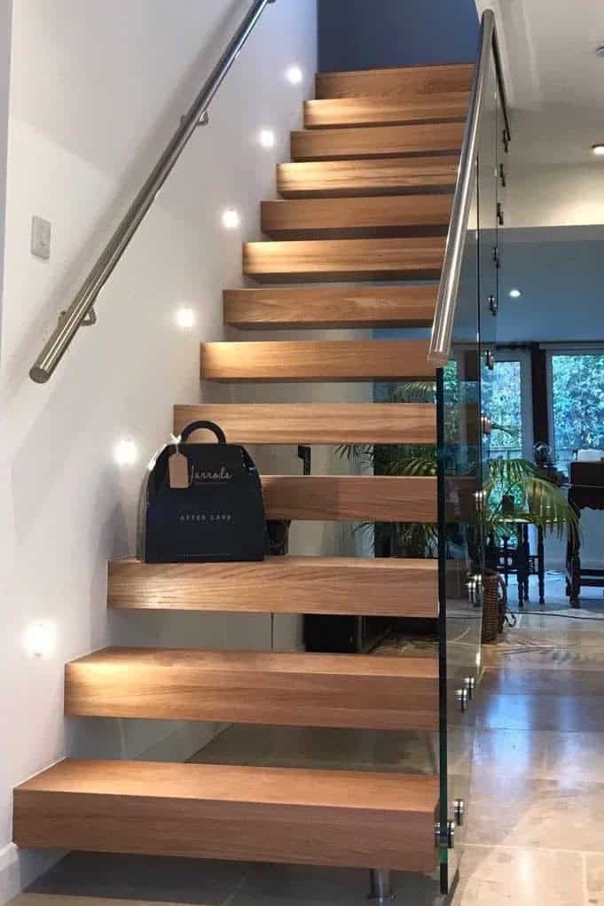 Straight oak open-riser staircase with a glass balustrade, metal handrails and bright recessed wall lights beside the treads.