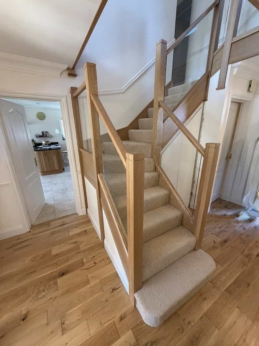 Beige carpeted turning staircase with oak-framed integrated glass balustrades, square posts and a rounded carpeted bottom step on a wooden floor.