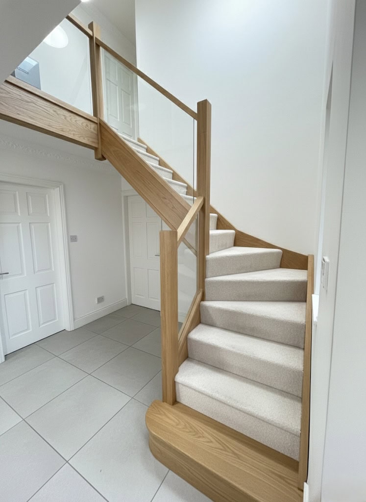Cream carpeted turning staircase with oak-framed glass balustrades, a rounded oak feature step and a matching upper landing in a bright tiled hall.