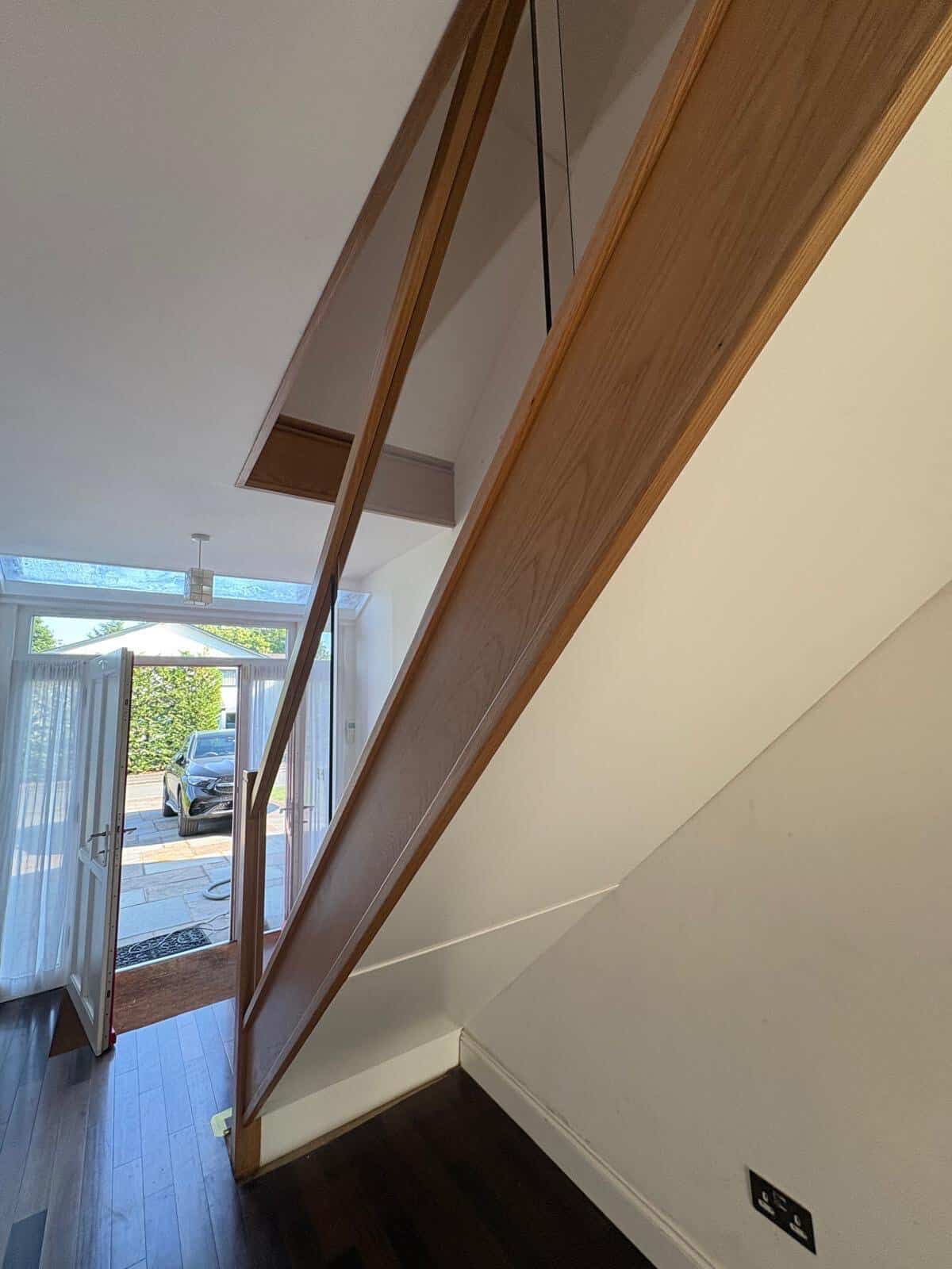 Side view of an oak staircase with clear glass panels integrated into the wooden rails above a white understair wall and dark hallway floor.