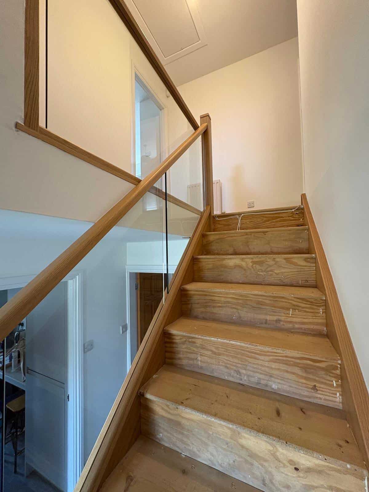 View up a staircase during renovation with unfinished wooden treads and new oak-framed integrated glass balustrades continuing around the landing.