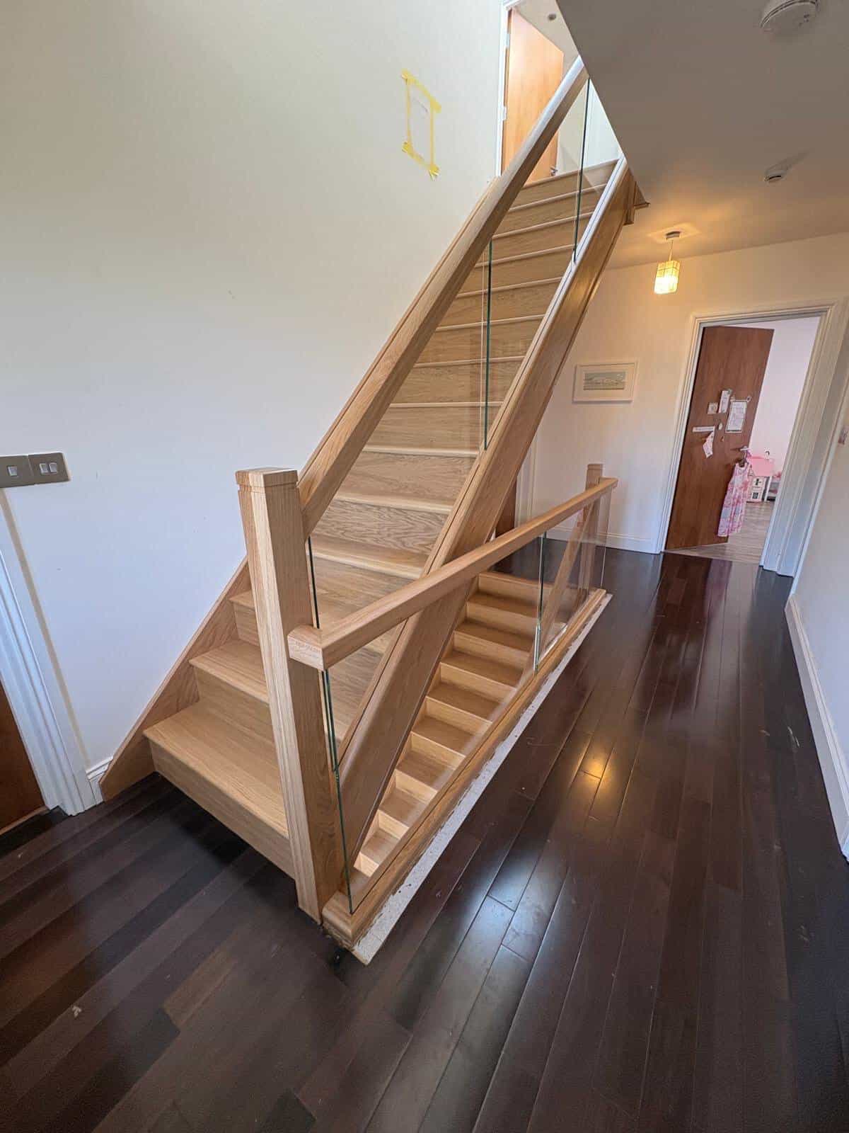 Pale oak staircase with integrated glass panels and a matching stairwell balustrade beside dark wooden flooring.