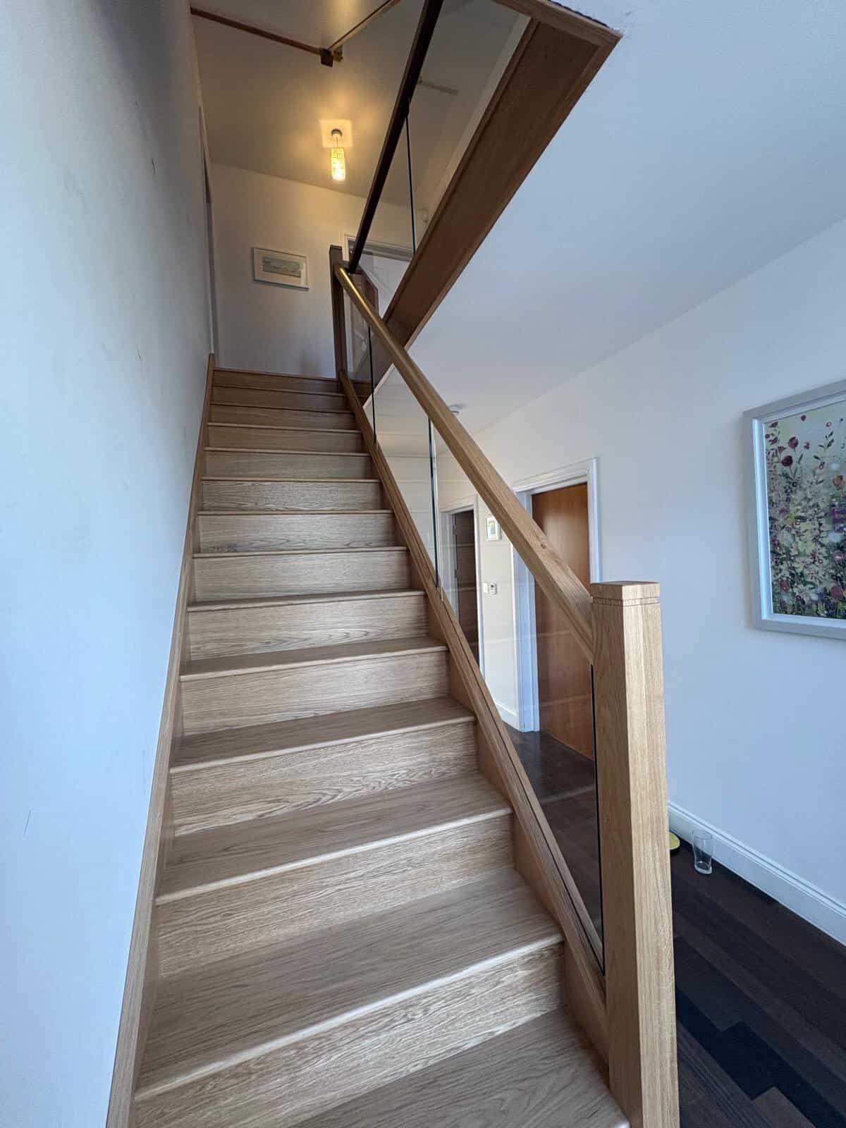 Straight pale oak staircase with integrated glass panels, square wooden posts and a matching glass-fronted upper landing in a white hallway.