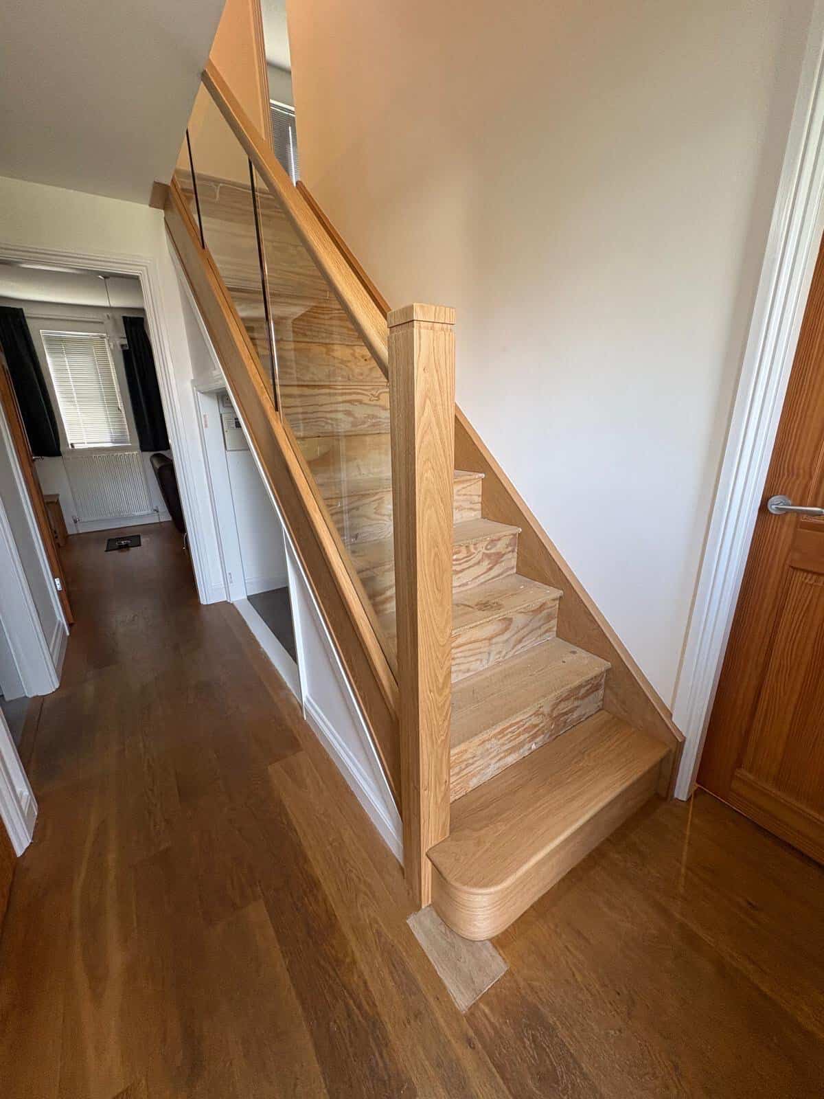 Oak staircase with prominent natural grain, integrated glass balustrades and a rounded feature step beside a wooden hallway door.
