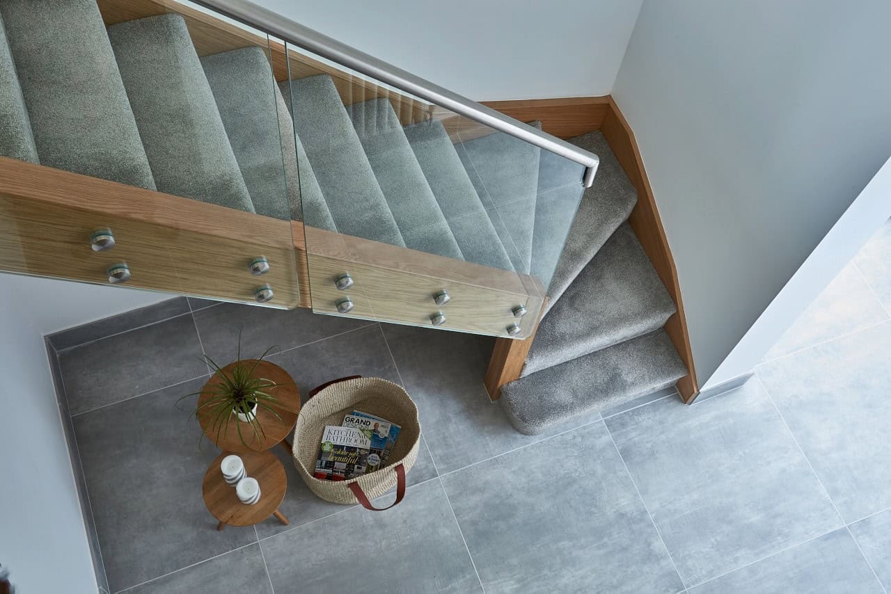 View down a renovated oak staircase with grey carpet, frameless glass balustrades and a metal handrail above a grey tiled hallway.