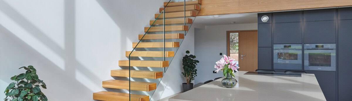 Floating oak staircase with open treads and a frameless glass balustrade above a contemporary kitchen with dark cabinets.