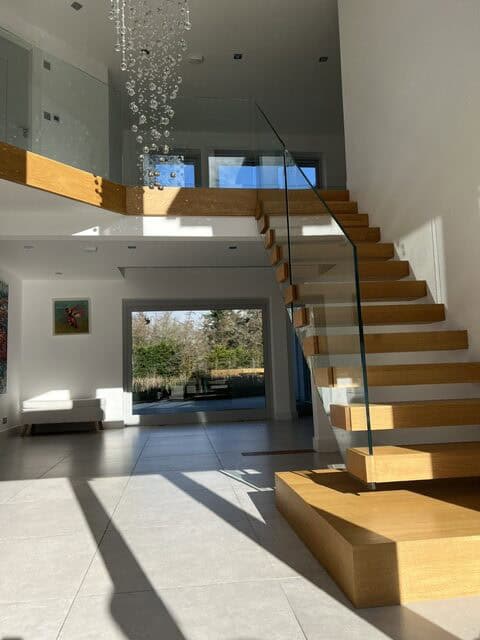 Floating oak staircase with frameless glass balustrades and a broad feature step beneath a chandelier in a sunlit double-height entrance hall.
