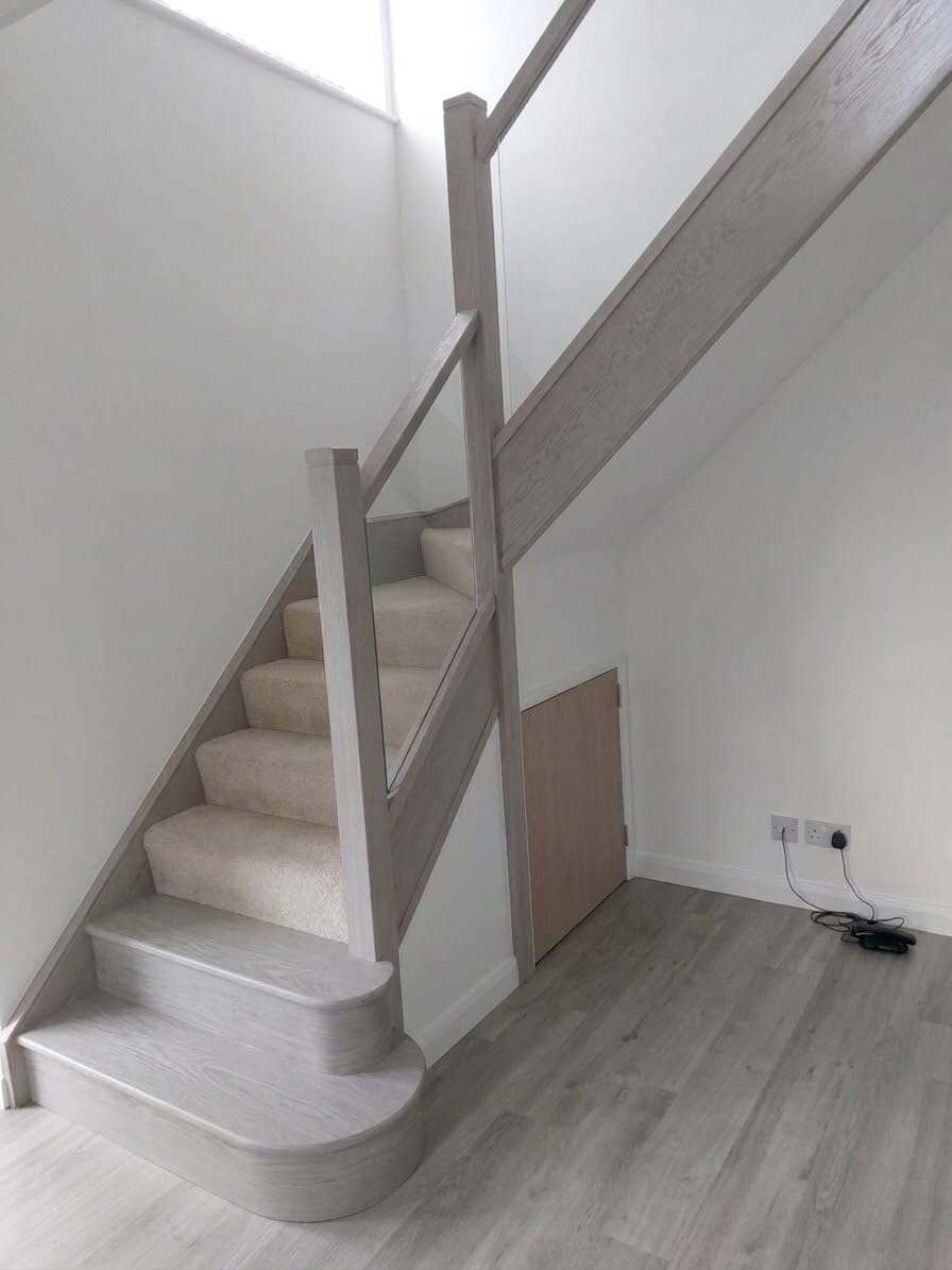 Pale grey-stained oak staircase with cream carpeted treads, integrated glass panels, square posts and two rounded wooden feature steps.