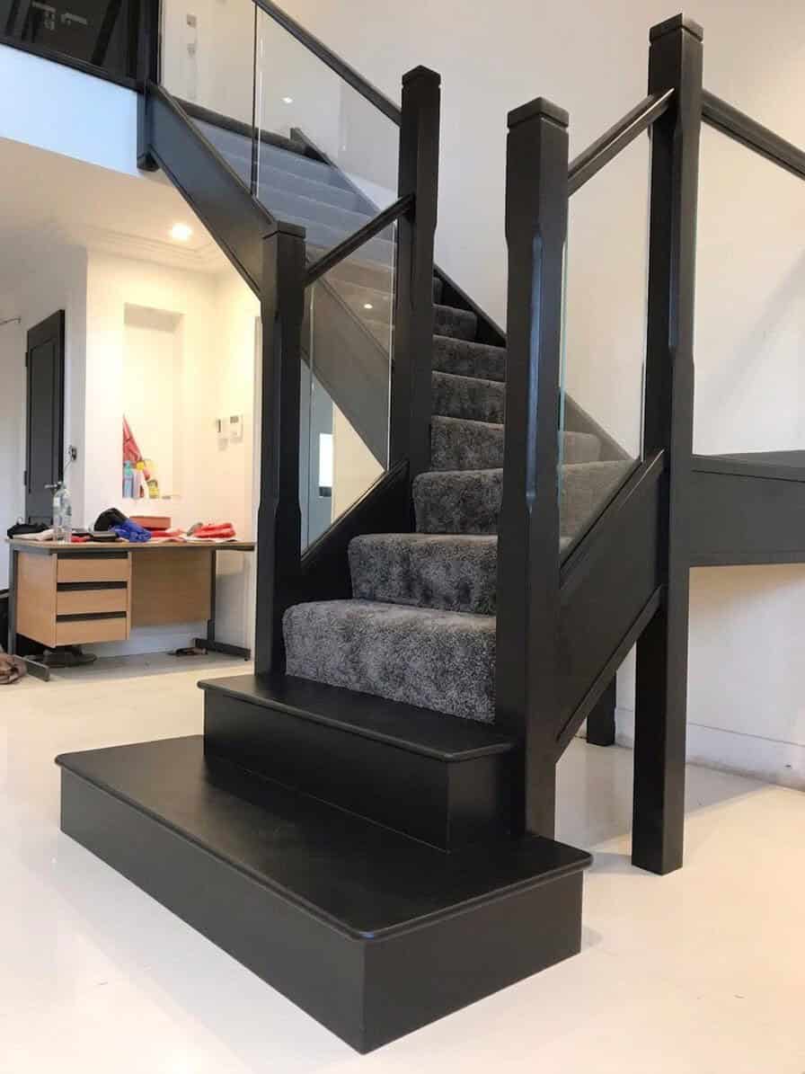 Black-stained oak staircase with grey carpeted treads, integrated glass balustrades, tall square newel posts and broad rectangular black feature steps.