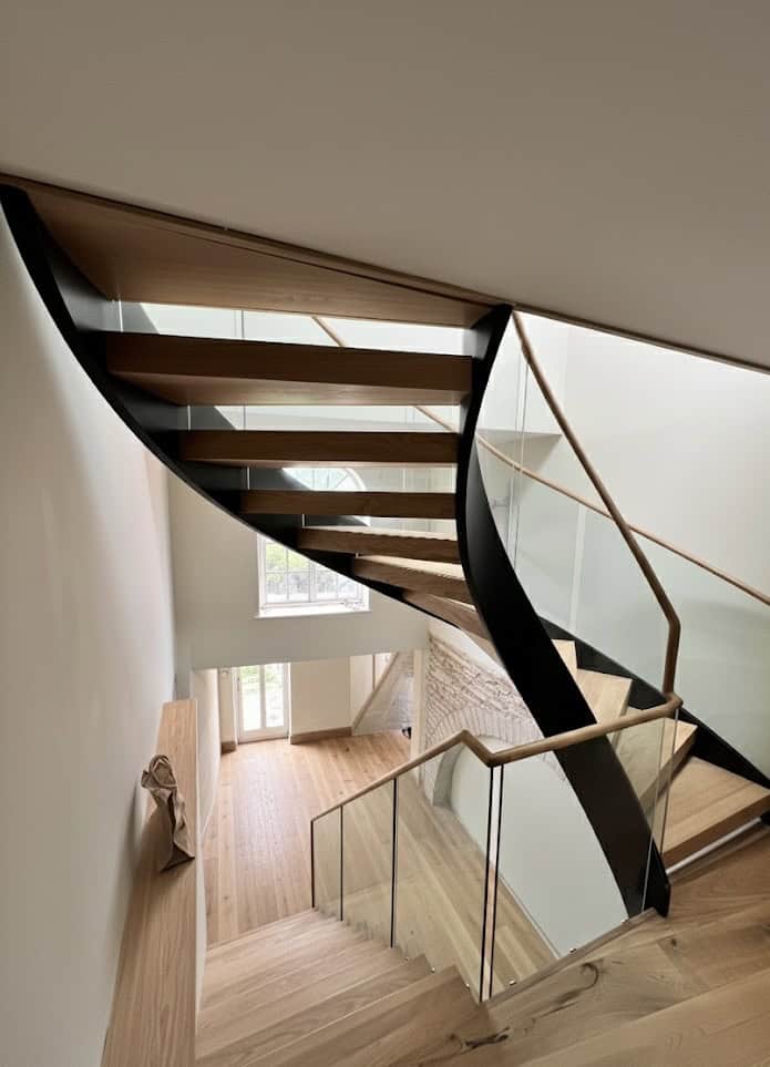 View down a curved open-riser wooden staircase with a black outer stringer, glass balustrades and wooden handrails.