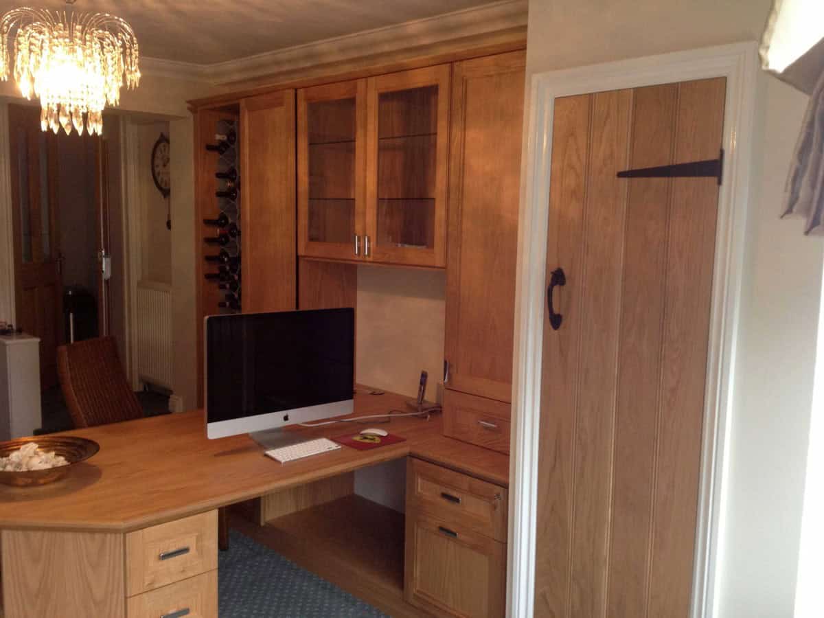 Bespoke oak home office with a curved desk, fitted drawers and cupboards, glazed upper cabinets and a vertical wine bottle rack beside a wooden door.