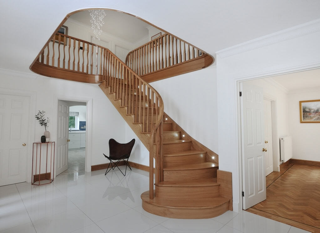 Sweeping oak staircase with turned wooden spindles, recessed step lights and a rounded bottom step beneath a curved gallery landing in a white hallway.