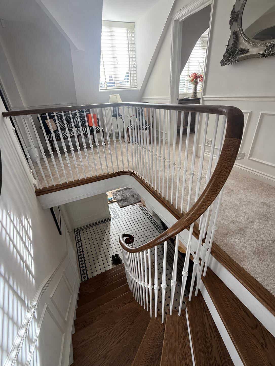 View down a dark wooden turning staircase with slender white metal spindles and a curved dark handrail around a carpeted upper landing.