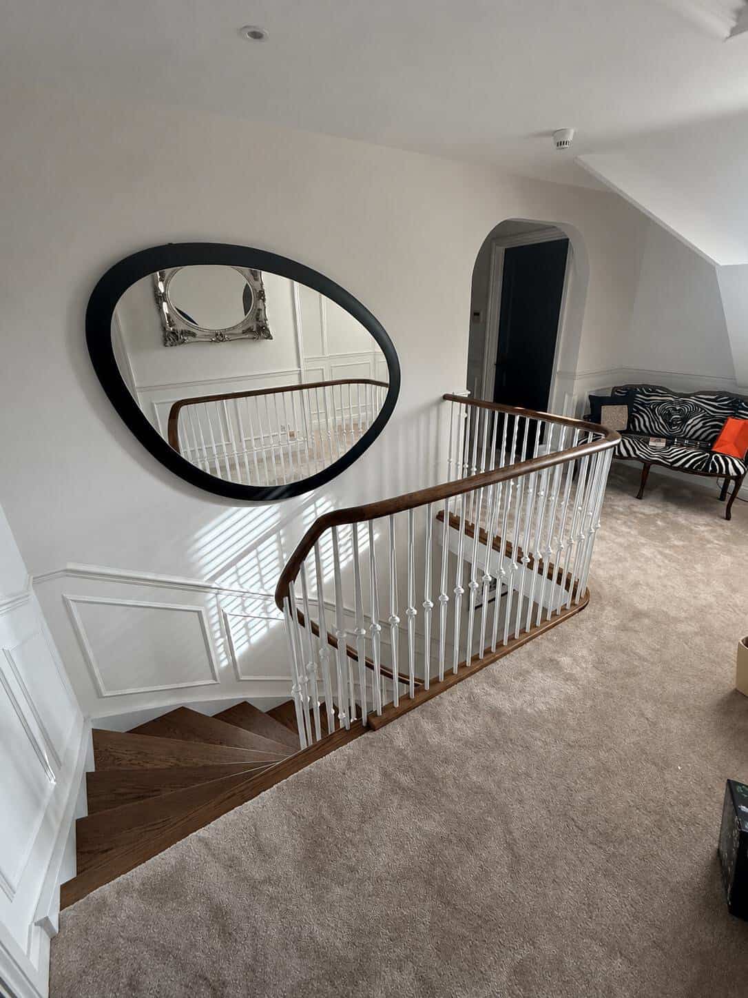 Carpeted upper landing with white decorative metal spindles and a curved dark wooden handrail around the stairwell beside a large round wall mirror.