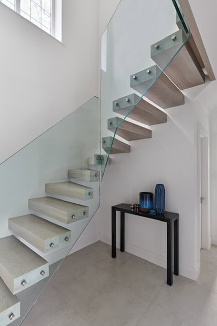 Pale wooden open-riser staircase with frameless glass balustrades and round metal fixings against white hallway walls.