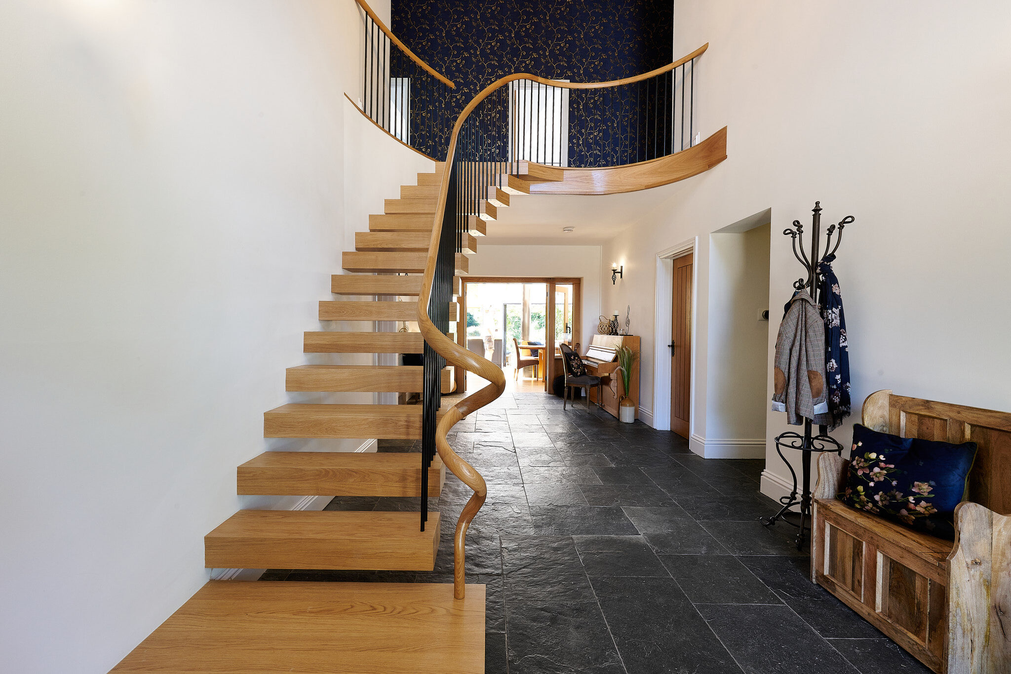 Floating oak staircase with black metal spindles and a flowing curved handrail leading to a matching gallery landing above dark stone flooring.
