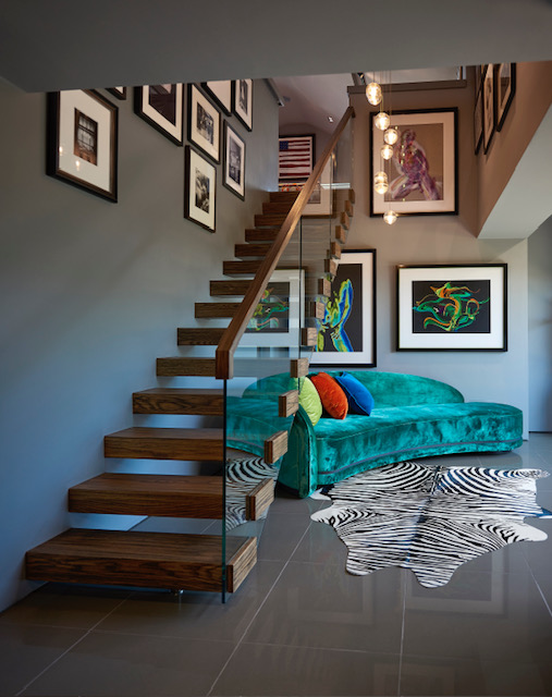 Dark wooden floating staircase with a glass balustrade and timber handrail beside a turquoise sofa and framed artwork.
