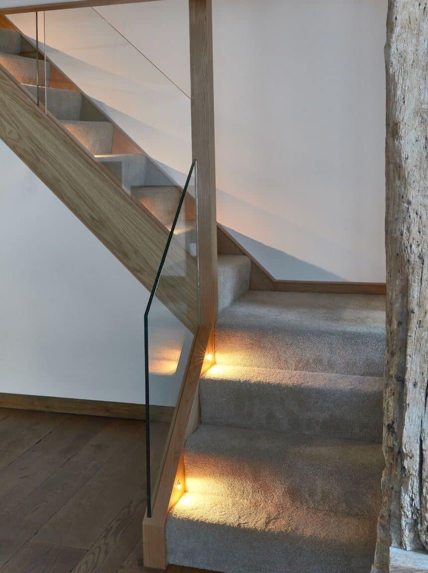 Turning grey carpeted staircase with oak framing, clear glass panels and warm recessed step lights beside an exposed stone wall.