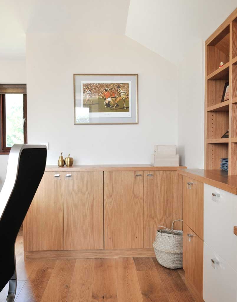 Fitted oak study cupboards with a continuous wooden countertop, open bookshelves and contrasting white drawers beside an office chair.