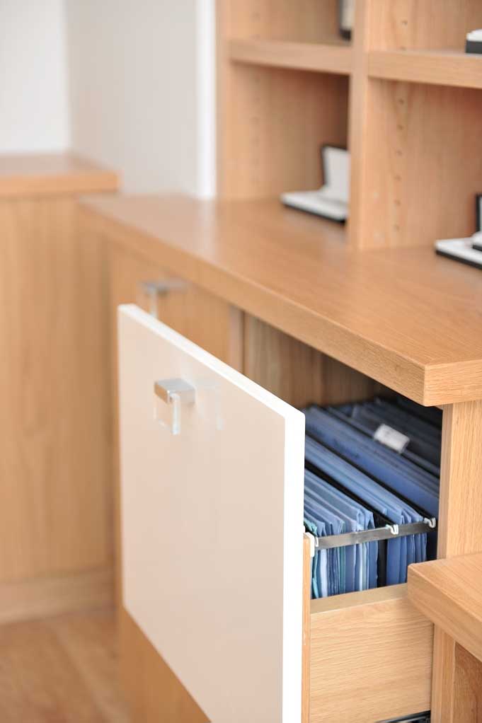 Open pull-out filing drawer with a cream front and hanging folders beneath an oak study countertop and open shelves.