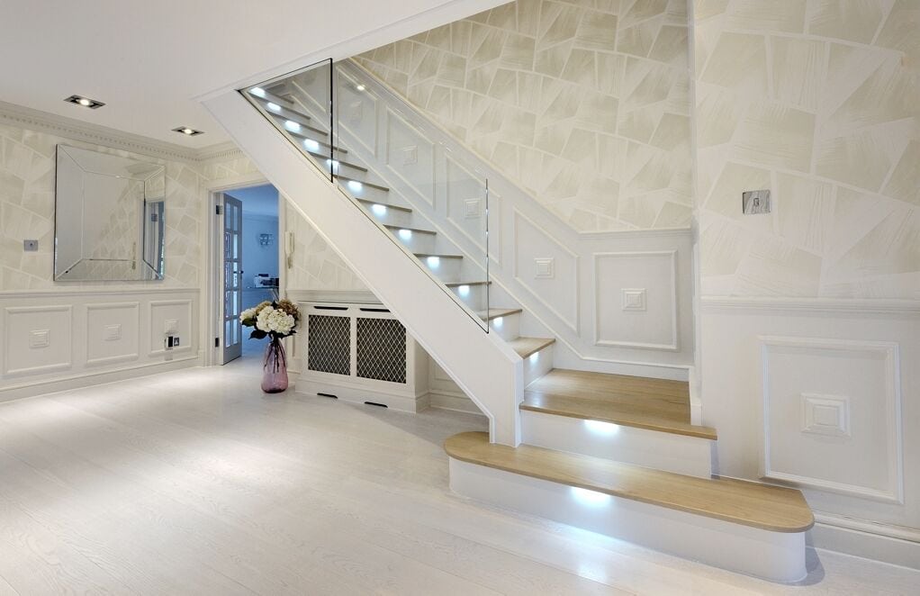White staircase with oak treads, recessed step lighting and clear glass balustrades above two broad rounded feature steps in a panelled hallway.