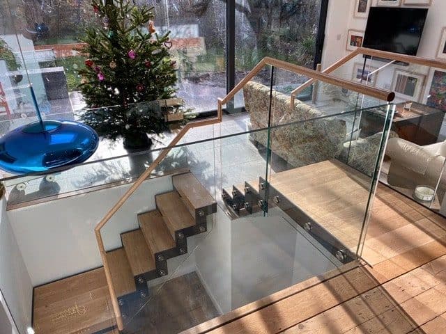 View down an oak staircase and landing with frameless glass balustrades and wooden handrails beside an open-plan living room.